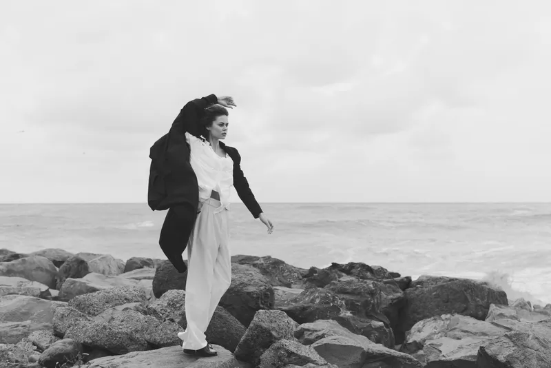 Woman walking on rocky coast in a flowing coat, wind swept, moody ocean backdrop, outdoor fashion in monochrome, dynamic movement across stones