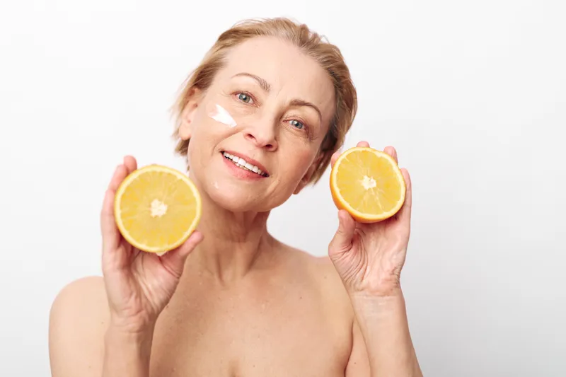 Mature woman with natural skin holds two halves of fresh orange, smiling gently. Facial cream applied on cheek, concept for skincare, wellness, natural beauty, and healthy lifestyle.