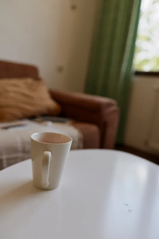 Cozy living room scene with a white ceramic mug on a smooth table, soft morning sunlight, relaxed mood, warm beige tones, cushions and drapes in the background, home lifestyle moment