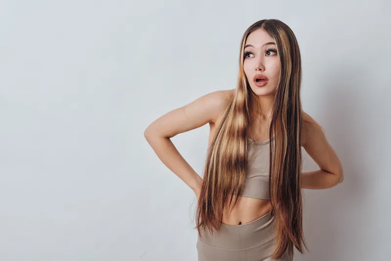 Young woman with long braided hair standing with hands on hips looking sideways with surprised expression on light background for fashion and beauty concept.