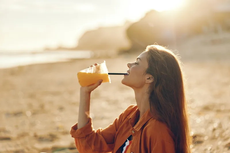 Woman drinking juice outdoors at sunrise, warm sunlight, seaside rocks background, orange drink in cup, health hydration lifestyle, summer wellness, calm candid moment, in fresh air