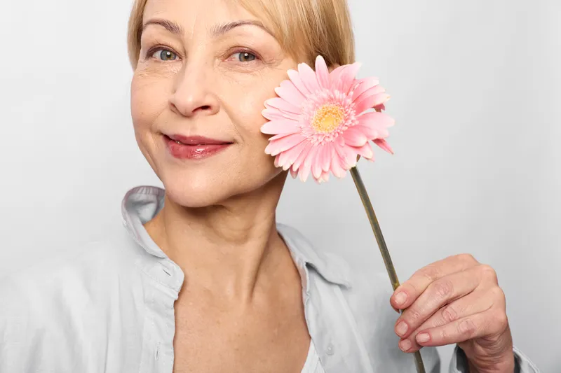 Woman portrait with smile holding pink flower near cheek, close up on white background, skincare and beauty concept, soft studio light, self care for wellness