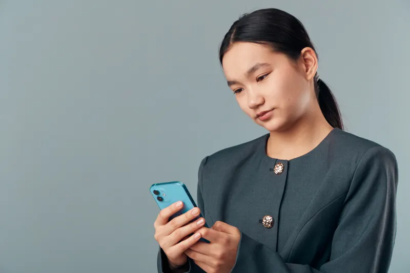 Young woman with long hair wearing a dark blazer looking at smartphone with focused expression on gray background. Female using mobile device for communication or information.