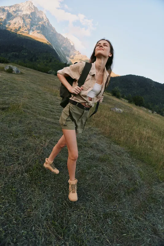 Young woman with backpack hikes through a mountain meadow, looking up at the sky with a sense of freedom and adventure. Sunlit scene with rugged mountains in the distance, a light breeze and natural