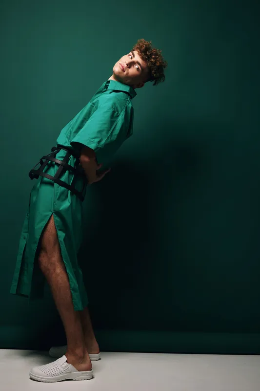 Young man with curly hair wearing green dress and white shoes posing against dark green background. Fashion model standing with hands behind back in studio with dramatic lighting.