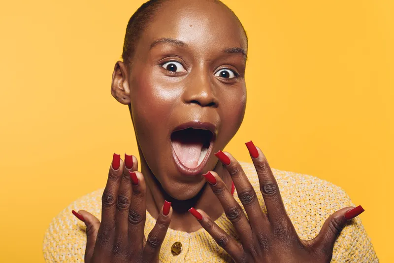 Surprised black woman with dark skin and short hair shows open mouth and red nails on yellow background. Concept of shock, excitement, beauty, and emotions in studio portrait.