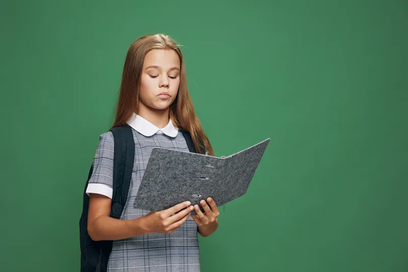 Girl student with backpack standing against green background reading open folder or notebook. Young schoolchild concentrated on study with focus preparing for lesson or homework indoors.