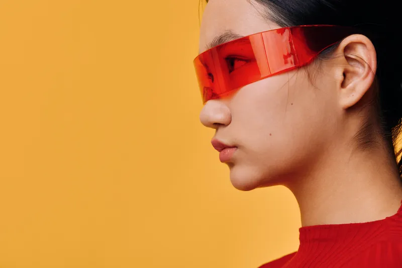 Profile of young woman with short black hair wearing red futuristic sunglasses and red top against solid orange background for fashion style and modern look.