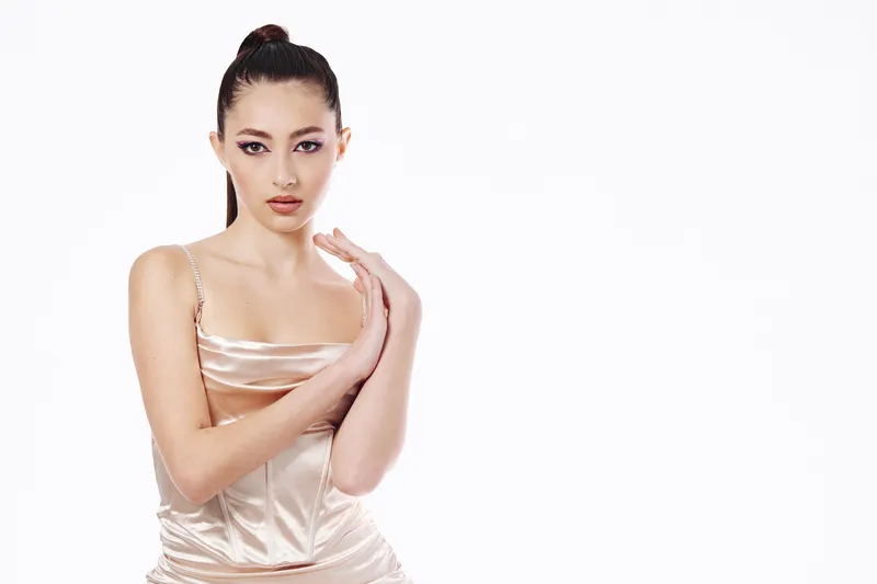 Young woman in a silky beige dress poses with folded hands against a white background. She has high-fashion makeup and sleek hairstyle, creating a beauty creative concept.