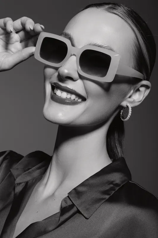Smiling Caucasian woman with pale skin wearing large sunglasses and hoop earrings in studio. Concept of fashion, beauty, and confidence.