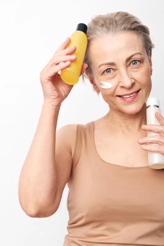 Mature woman with light skin and blue eyes applies cream on face holding two cosmetic bottles. Concept of skincare routine, natural beauty, anti-aging care, wellness, and self-care at home.