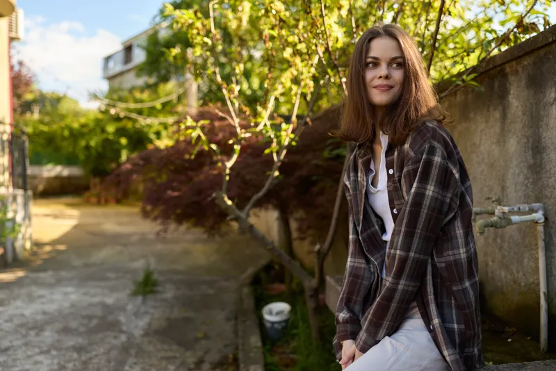 Woman lifestyle portrait outdoors in sunny garden wearing plaid shirt, casual white dress, relaxed pose, smiling expression, natural light, candid moment of daily style