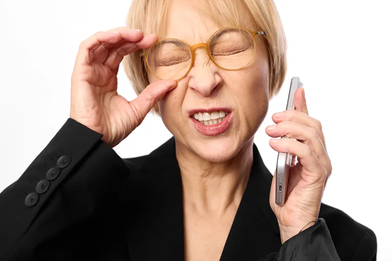Woman with glasses holding a phone, business portrait against white background, expressing stress and frustration, possible headache, studio lighting, closeup