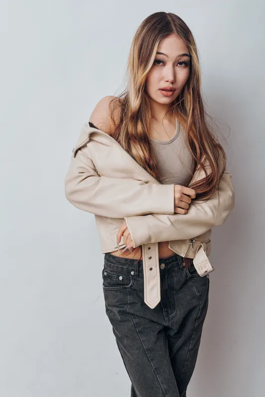 Young woman with long light hair wearing beige jacket and grey tank top posing against white wall with casual expression in studio portrait. Fashion, style and beauty concept.