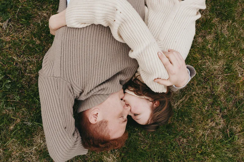 An affectionate couple lies on grass in cozy sweaters, sharing a close moment outdoors. A relaxed lifestyle portrait highlighting connection, warmth, and simple happiness in a natural setting.