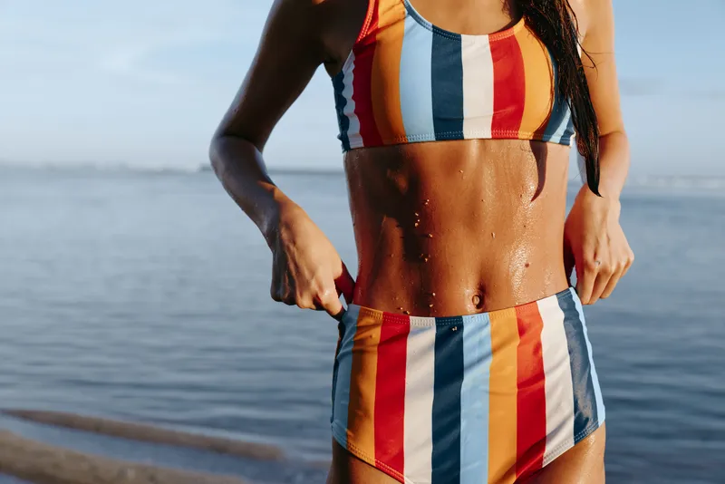 Woman beach bikini ocean striped sunny summer confidence adjusting swimsuit near ocean horizon with warm sunlight saltwater sparkle on skin carefree summer lifestyle travel relaxation moment