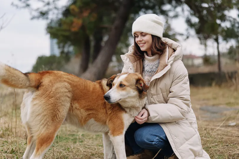 Woman in a warm winter coat kneels beside a friendly dog in a park, sharing a calm moment outdoors among trees and soft daylight, showing companionship and trust during a peaceful outdoor stroll