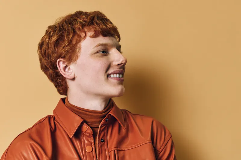Young man with red hair and fair skin wearing brown jacket looking sideways on beige background. Portrait of confident male model with casual style and wavy hair, smiling and relaxed.
