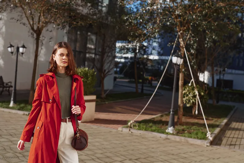 Young woman with red hair wearing a long red coat walks confidently on a sunny urban street with trees and modern buildings in the background.
