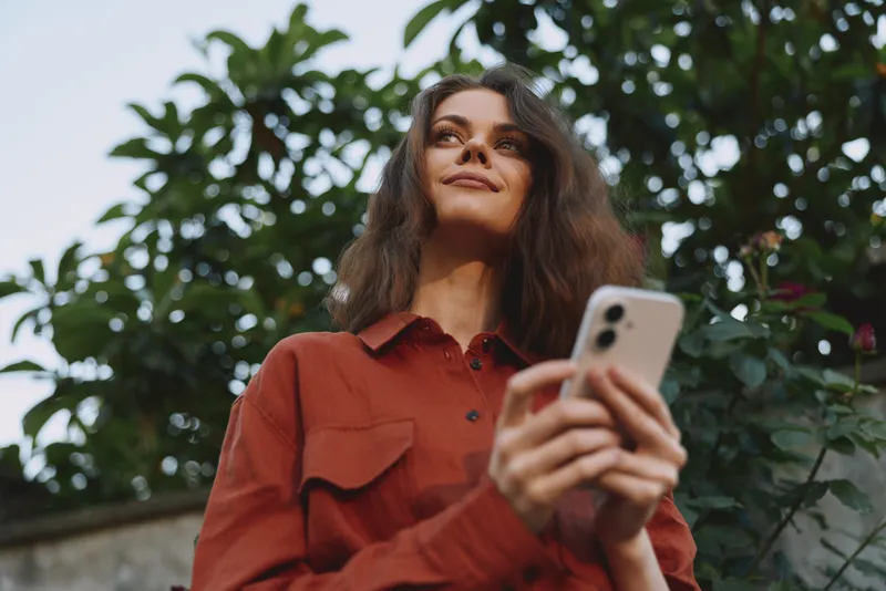 Young woman with long hair wearing a rust shirt stands outdoors among greenery, looking up with a relaxed smile while holding a smartphone, breeze lifting her hair, soft light and natural colors