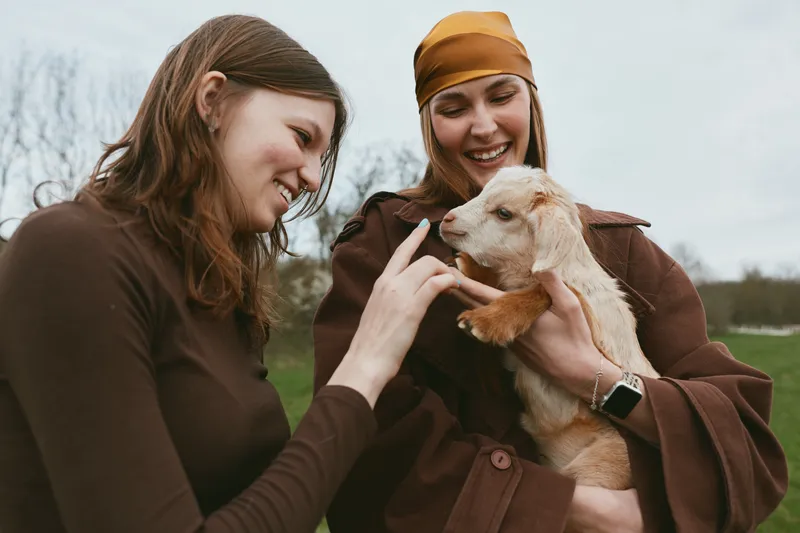 Two young women enjoy a moment outdoors as one holds a baby goat while the other gently touches its nose, both smiling and dressed in warm brown clothing on a cloudy day