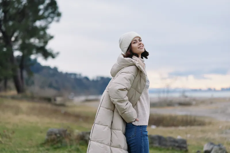 Winter outdoor portrait of a woman in a long beige coat, scarf and beanie, standing in a windy coastal field with trees and distant horizon