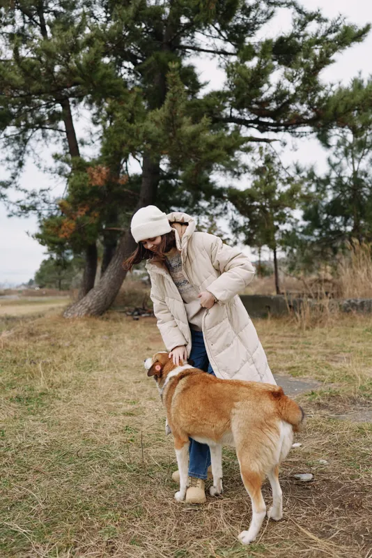 Woman in a warm coat and beanie interacts with a brown dog in an open park, trees in the distance, capturing a calm outdoor moment and simple companionship.