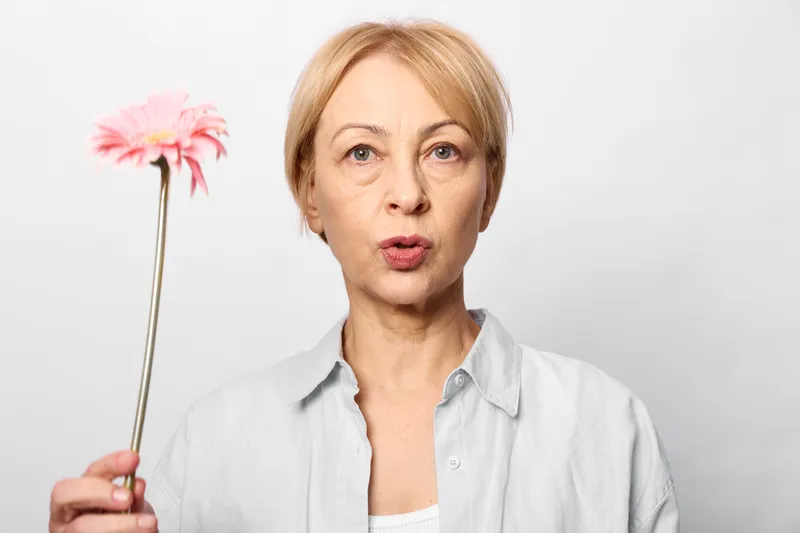 Woman holding a pink flower in a portrait on a clean white background, surprised expression. Soft studio lighting supports skincare, beauty, and spring freshness.