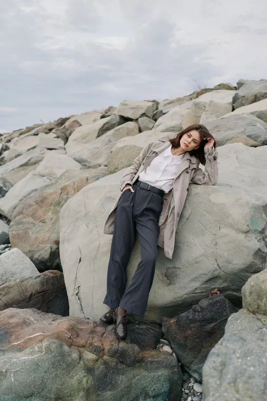 Young woman in stylish outfit leans on large rocks near the coast. Casual fashion, outdoor lifestyle, and natural scenery create a relaxed and modern mood.