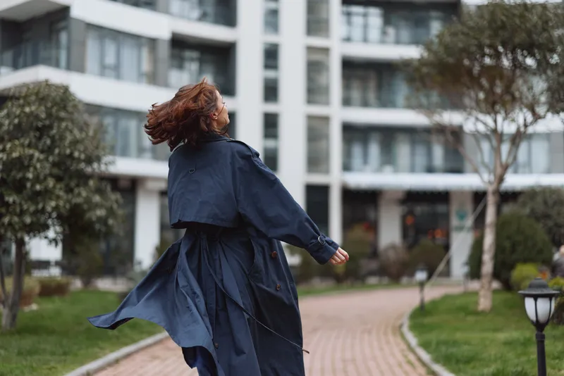 Young woman with curly hair wearing a flowing dark trench coat spins joyfully on a paved path near modern apartment buildings on a cloudy day.