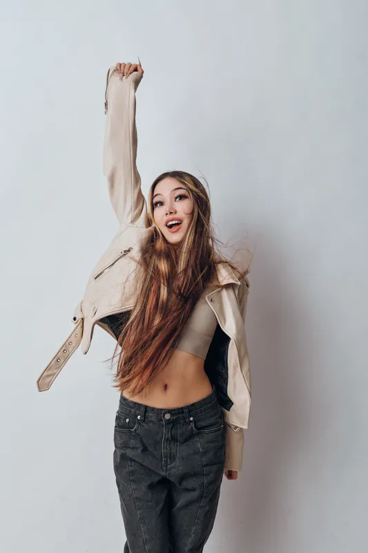 Young woman in casual clothing posing happily with raised arm against plain background, smiling and displaying energy and confidence in studio setting.