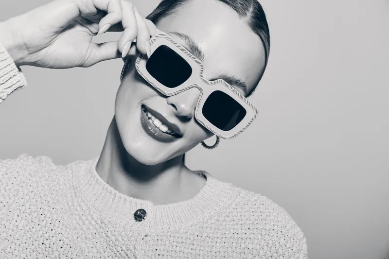 Portrait of a smiling young white woman with fair skin wearing large sunglasses and a knitted sweater in a bright studio. Concept for fashion, style, and accessories.