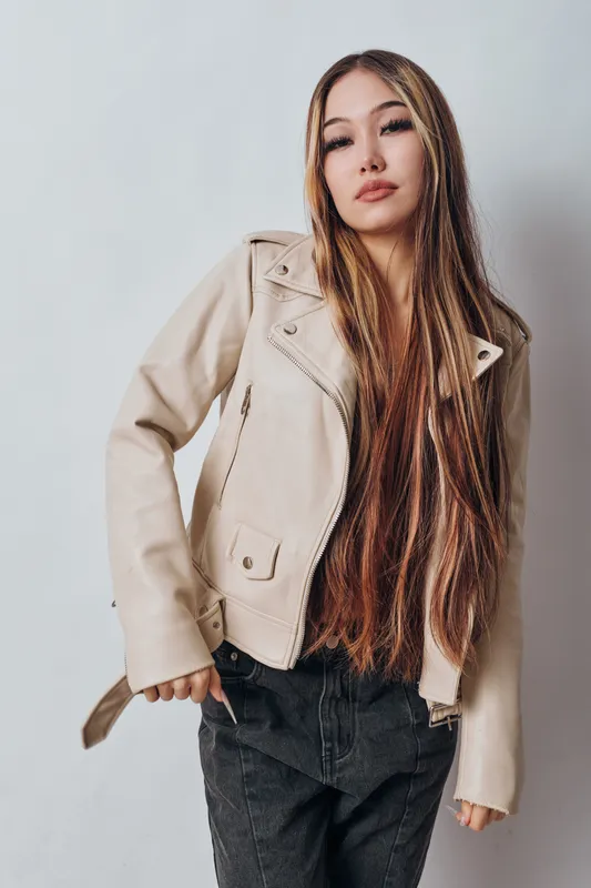 Young woman with long hair wearing a beige jacket and black pants posing confidently in front of a plain white background with hands in pockets and relaxed expression.
