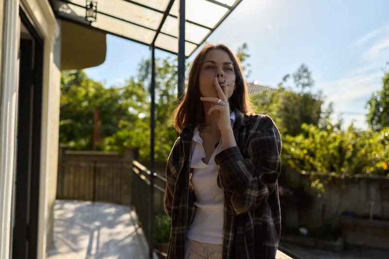 A stylish woman stands outdoors in a plaid jacket, smiling softly as she touches her lips. A sunny patio scene conveys relaxed, everyday life and casual fashion moments