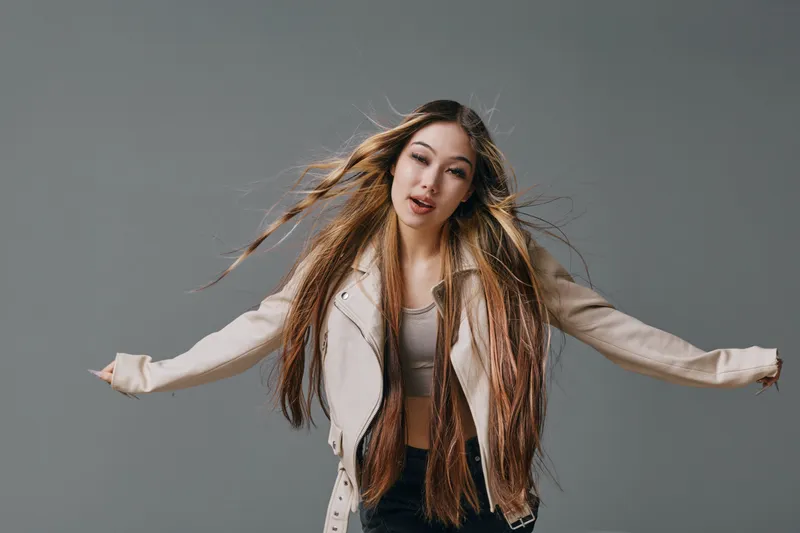 Young woman with long hair wearing light jacket and crop top posing with arms outstretched on gray background. Fashion model showing movement and casual style indoors.