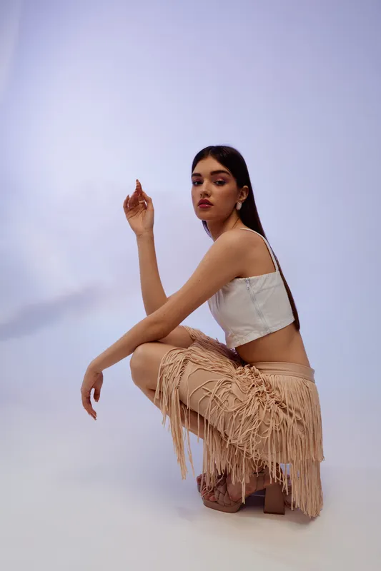 Young woman in a white top and fringed skirt poses confidently against a soft background. Concept of beauty, fashion, and style in modern portrait photography.