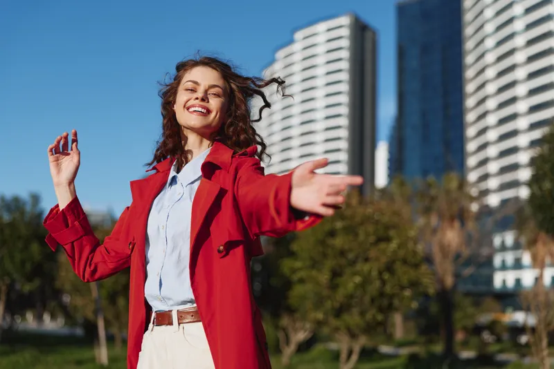 A young woman with curly hair wearing a red coat joyfully reaches out while standing outdoors near modern buildings on a sunny day.