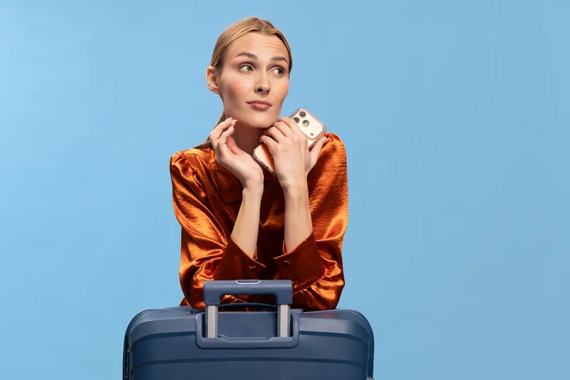 Young woman in orange satin blouse holds smartphone and leans on blue suitcase, looking away. Concept for travel, vacation, planning, and modern lifestyle with casual fashion.