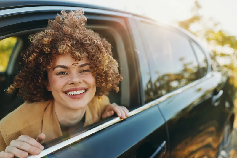 Happy woman with curly hair leaning out of a car window, smiling warmly, casual outfit, sunny outdoor scene, natural light, candid lifestyle moment, vibrant retro vibe