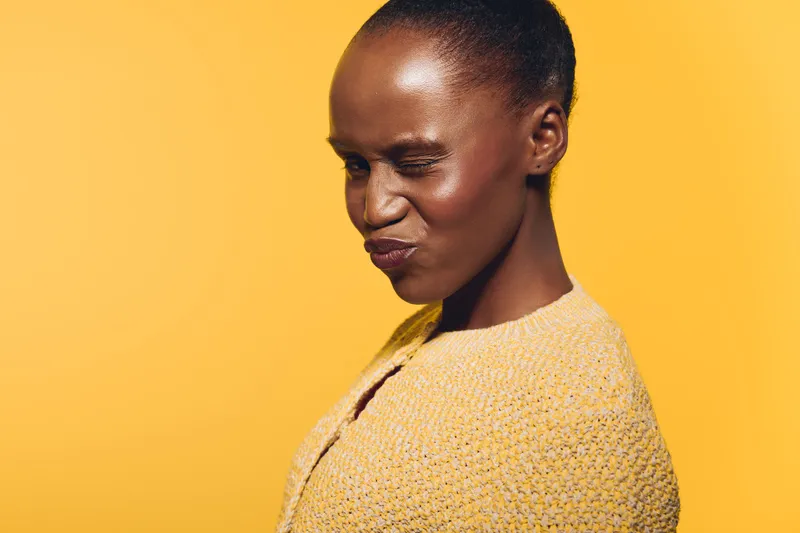 Portrait of a young black African woman with dark skin wearing a yellow knitted sweater on a yellow background. She has a playful expression with eyes closed and lips pursed.