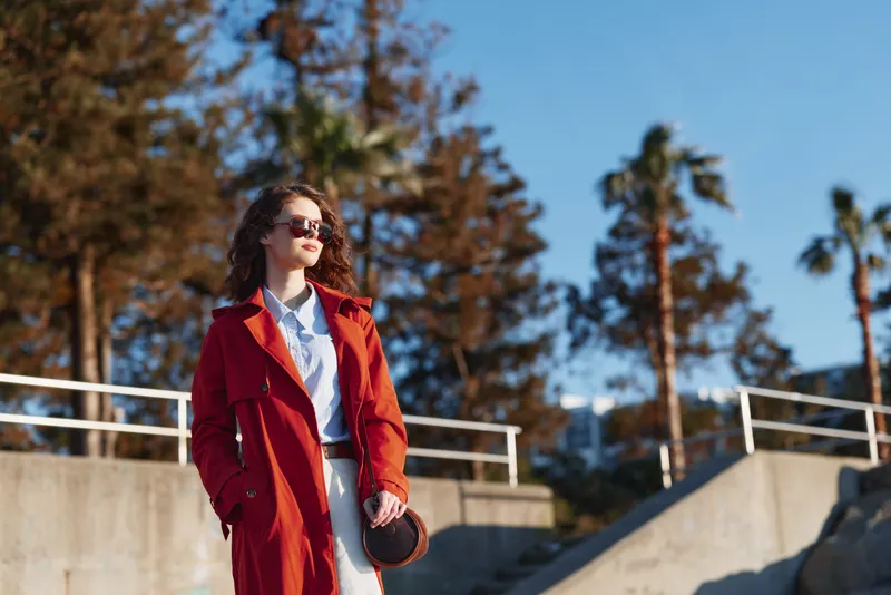 Young woman wearing a red coat and sunglasses walks confidently along a sunny urban street with palm trees and modern buildings in the background.