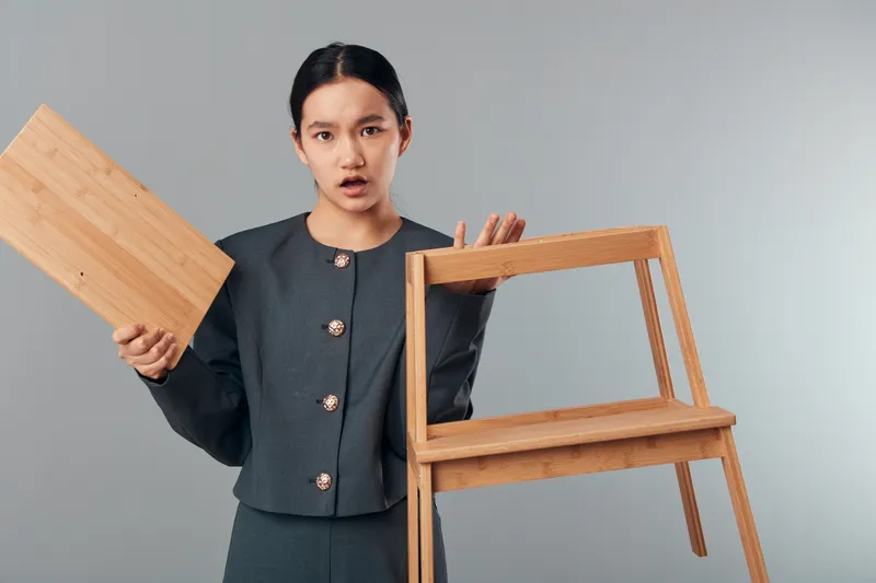 Young woman holding a broken wooden chair and a detached piece of chair in studio. Repair, damage, furniture and expression of surprise or confusion on gray background.