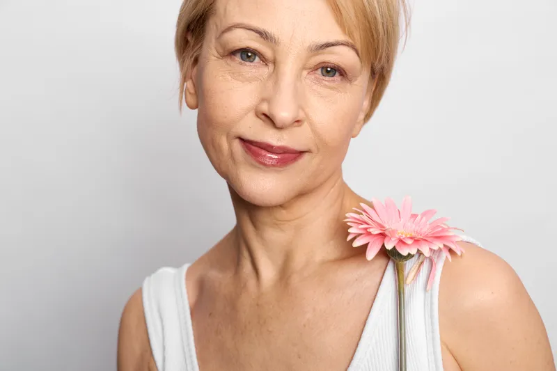 Senior woman portrait closeup headshot smiling holding pink flower on gray background soft daylight minimal studio look gentle expression with calm wellness mood.