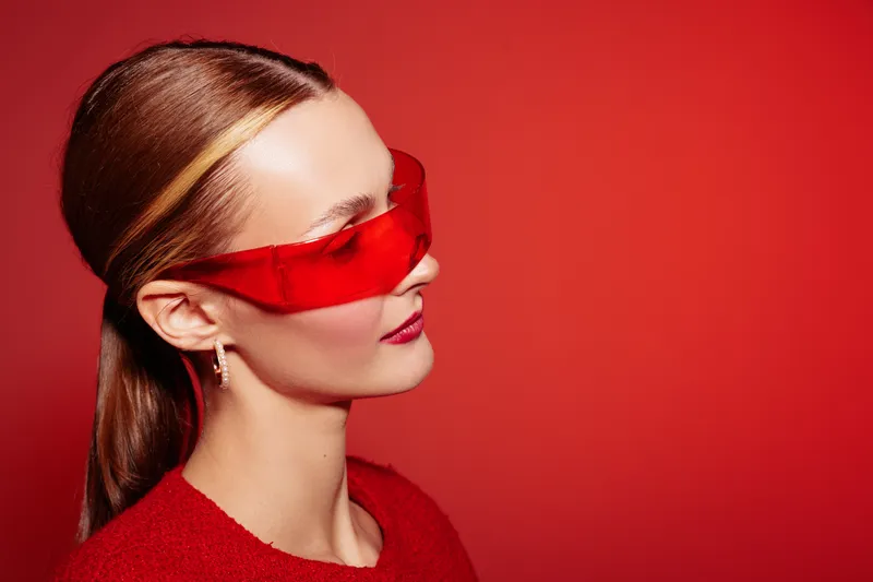 Young woman with red sunglasses and red sweater poses against red background. Side profile with sleek hair and bold makeup creates modern and stylish fashion concept.