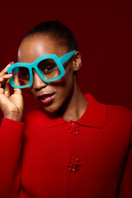Portrait of a confident dark skinned African woman wearing oversized blue glasses and a red sweater against a red background. Concept of fashion, style, and bold accessories.