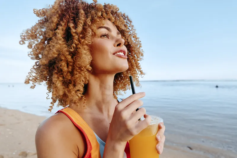 Woman with curly hair on beach, drinking water from bottle, smiling in warm sunlight, relaxed posture, summer outdoors near calm sea, clear sky, natural colors