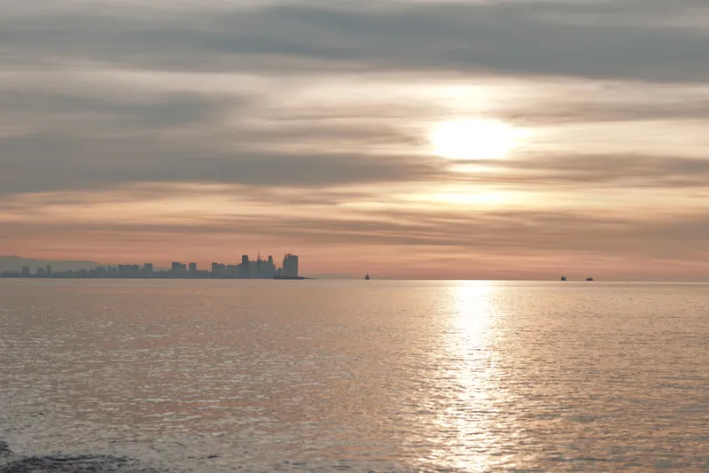 Sunset over calm water with a distant city silhouette, warm reflections on the sea and a tranquil horizon under soft clouds, creating a serene coastal scene and peaceful evening mood.
