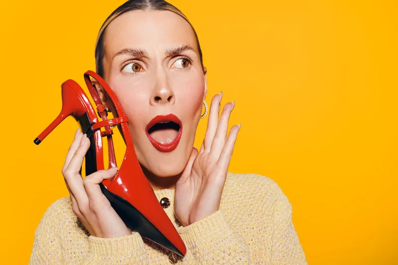 Surprised woman with red lipstick holds red high heel shoe near face on bright yellow background. Concept of fashion, style, beauty, and shopping excitement with vivid colors and expression.