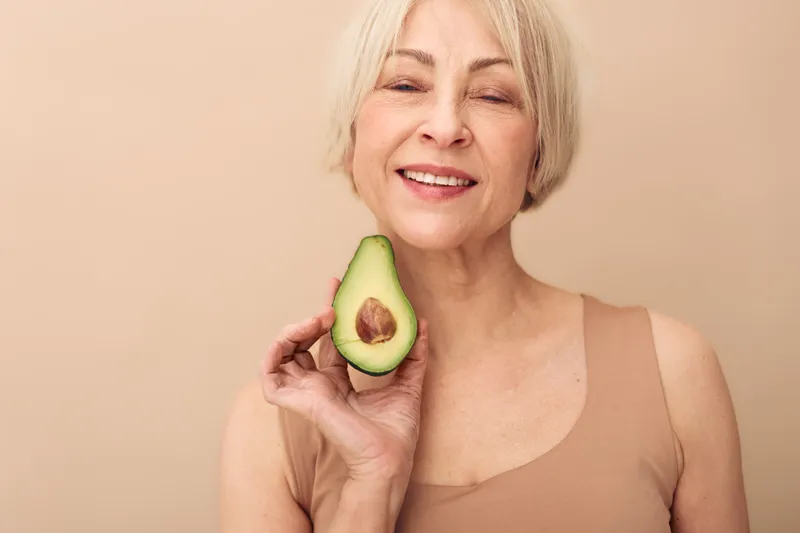 Smiling mature woman with short blonde hair holds half of fresh avocado near face on beige background. Concept of healthy lifestyle, nutrition, beauty, and natural skincare for older adults.