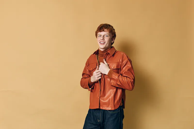Young man with red hair smiling and buttoning orange leather jacket standing against beige background casual fashion style and confident pose.